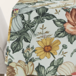 Tischdecke mit detailreichem Blumenmuster im Vintage-Stil, zeigt gelbe, rosa und cremefarbene Blüten sowie grüne Blätter auf hellem Hintergrund.